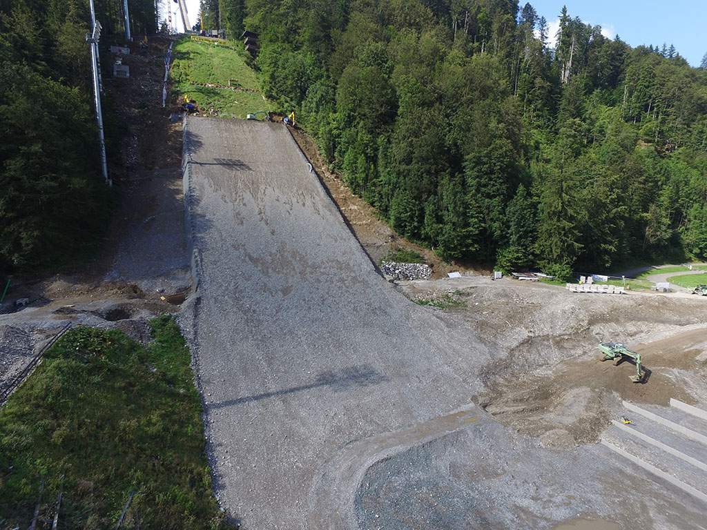 Aufnahme von einer Drohne;  Oben im Hang sieht man den gesteuerten Bagger;  Links und rechts davon je ein Schreitbagger, mit denen die Aushubarbeiten durchgeführt wurden.  Rechts unten ist ein weiterer Dual-GNSS-Bagger zu sehen (Liebherr 926), mit dem Er